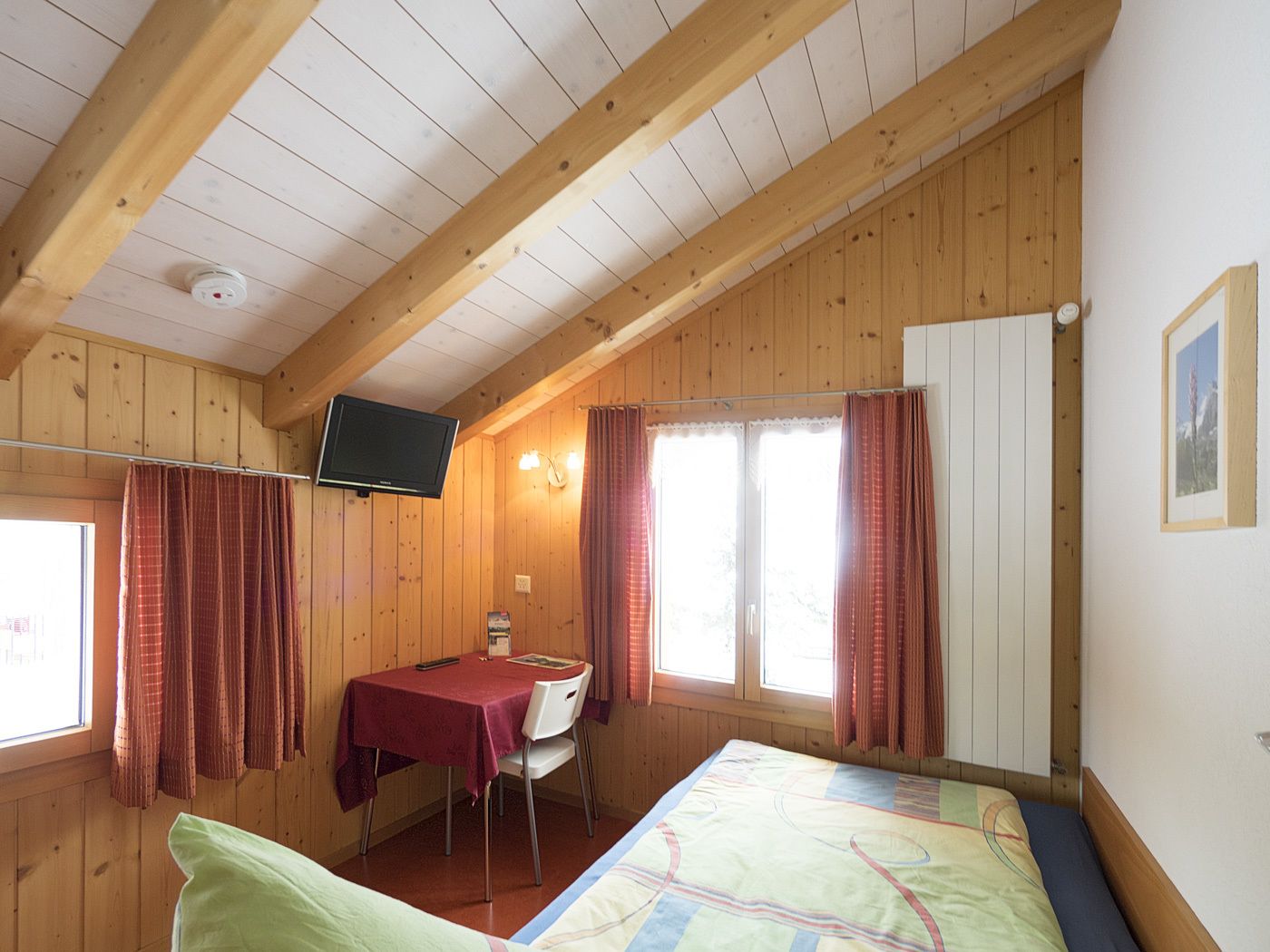 A cozy room with wooden walls, a bed, a table with a chair, a TV, and a window with curtains.
