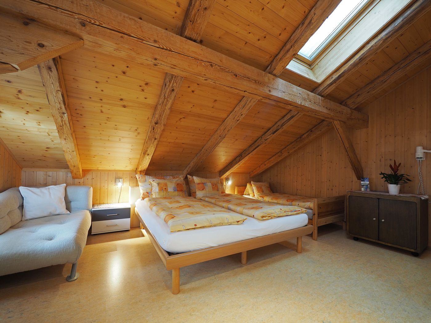 A cozy attic bedroom with a wooden sloped ceiling, a large bed, a sofa, and a cabinet.
