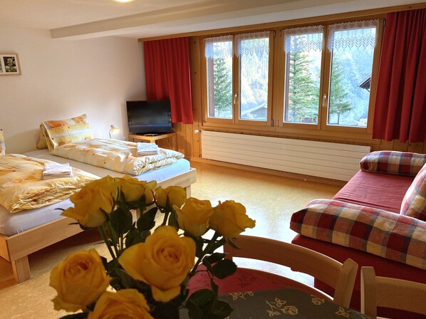 A cozy room with a bed, a sofa, and a TV. There are windows with curtains and a view of trees outside. - Esthers Guesthouse (Gimmelwald)