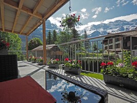 Un espacio al aire libre cubierto con una mesa de vidrio que refleja el cielo y las montañas, rodeado de plantas en macetas y una pérgola de madera.