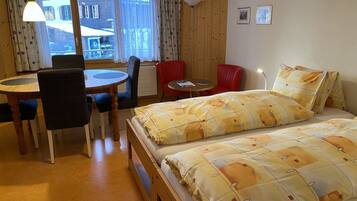 Una habitación con una cama, una mesa de comedor con sillas, una mesita redonda y una ventana con cortinas.