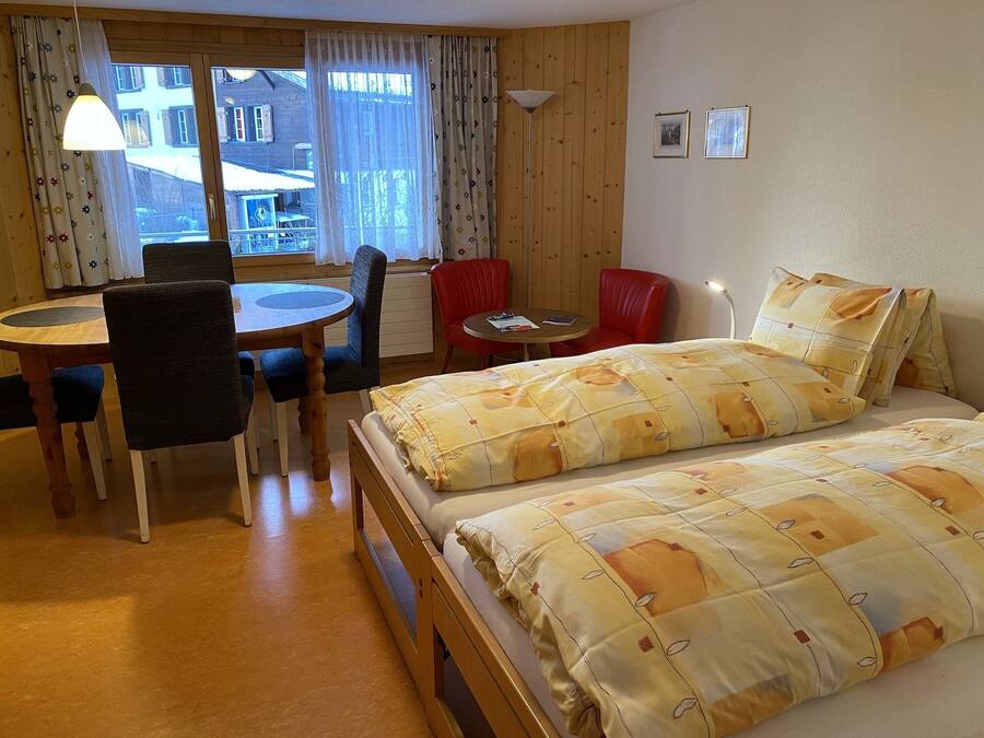 Una habitación con una cama, una mesa de comedor con sillas, una mesita redonda y una ventana con cortinas.