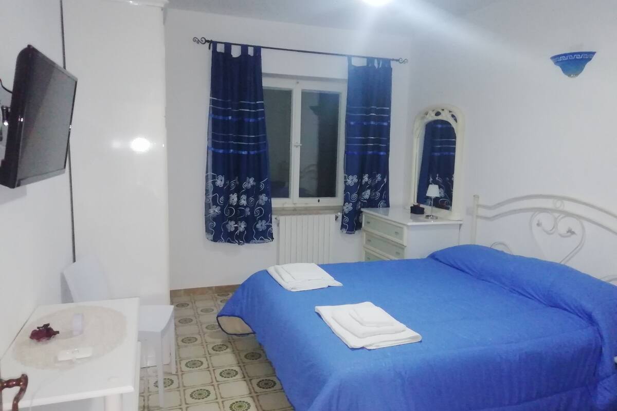 A bedroom with a bed, a small bedside table, a mirror, and a window with curtains.