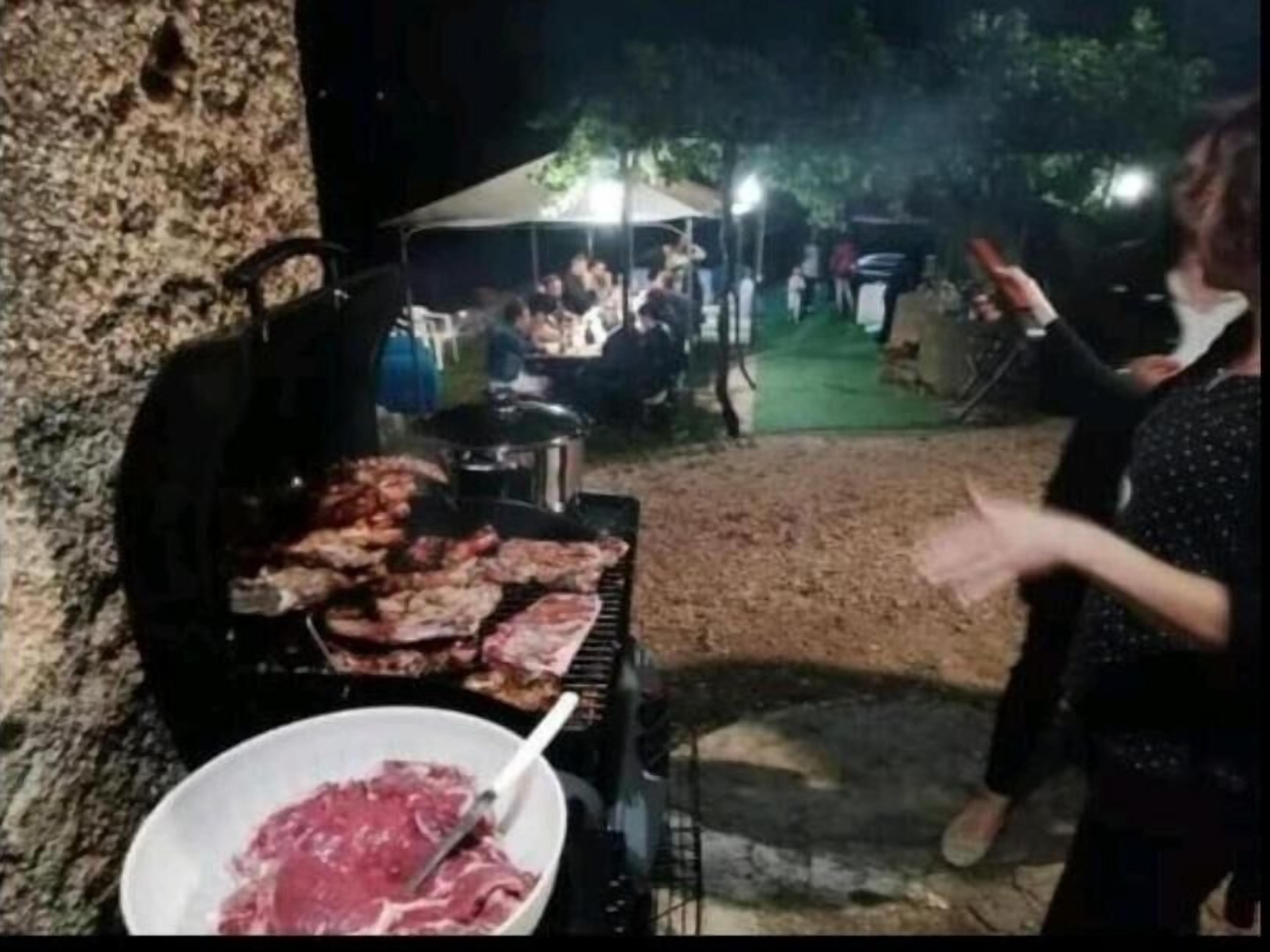 An outdoor barbecue with meat being cooked on a grill, a bowl of raw meat, and people in the background.