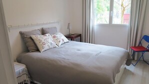 A bedroom with a bed, a nightstand, a lamp, a chair, and a window with curtains. - 004 - Relaxation and rest in this house 2 steps from the ocean (SOULAC SUR MER)
