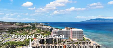 A high-rise hotel with multiple floors, located near a beach with clear blue waters and a mountainous backdrop.