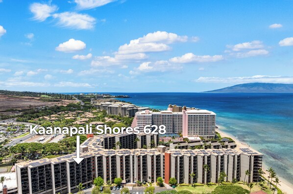 Un grand hôtel à plusieurs étages, situé près d’une plage aux eaux d’un bleu limpide, avec une chaîne de montagnes en toile de fond.