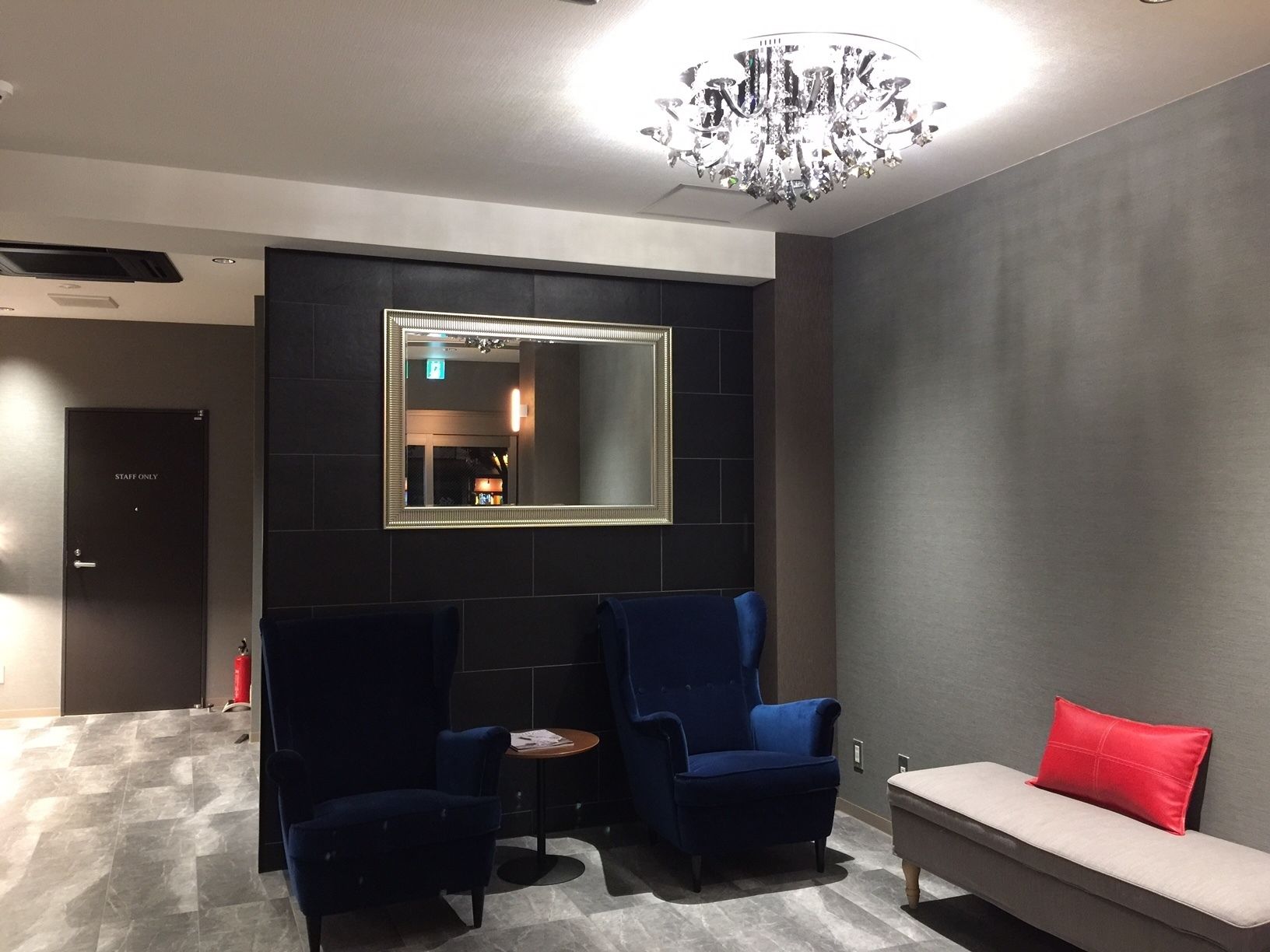 A modern lounge area with a large mirror, two blue armchairs, a small round table, and a sofa with a red pillow.