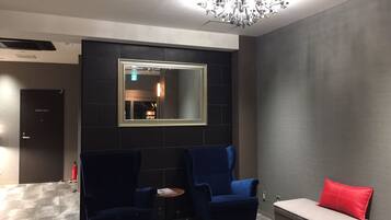 A modern lounge area with a large mirror, two blue armchairs, a small round table, and a sofa with a red pillow.