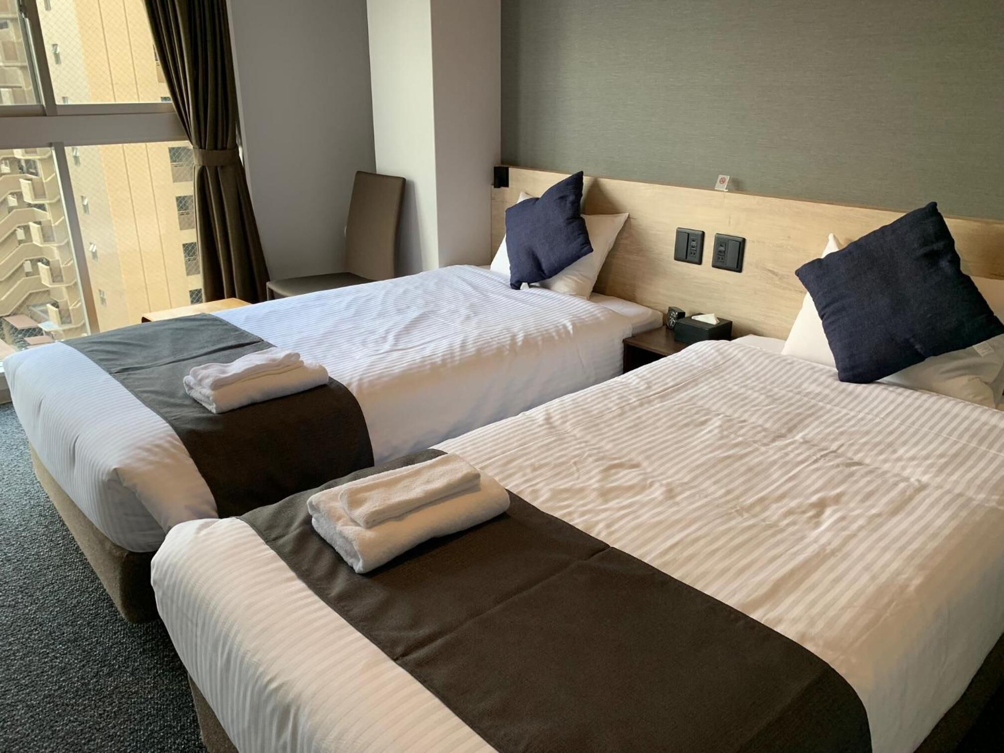 A hotel room with two beds, each with a pillow and a folded towel. There is a nightstand with a lamp and a power outlet.