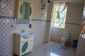 A bathroom with patterned walls, a white sink, a mirror, a toilet, and a window with curtains. - NP Plaza Hotel (Akosombo)