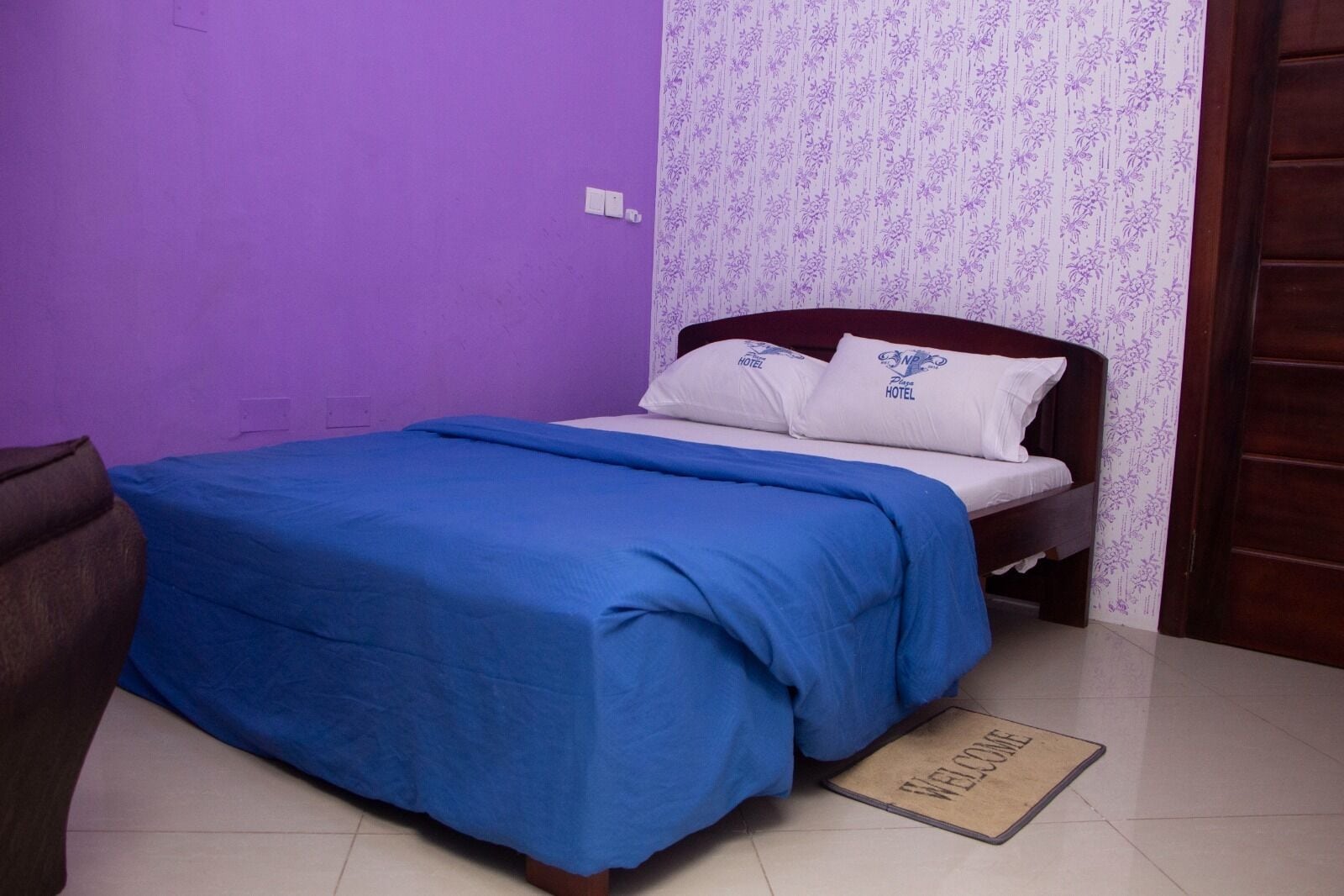 Una cama pequeña y bien hecha, con una colcha azul y almohadas blancas, una mesita de noche de madera y un tapete de bienvenida.