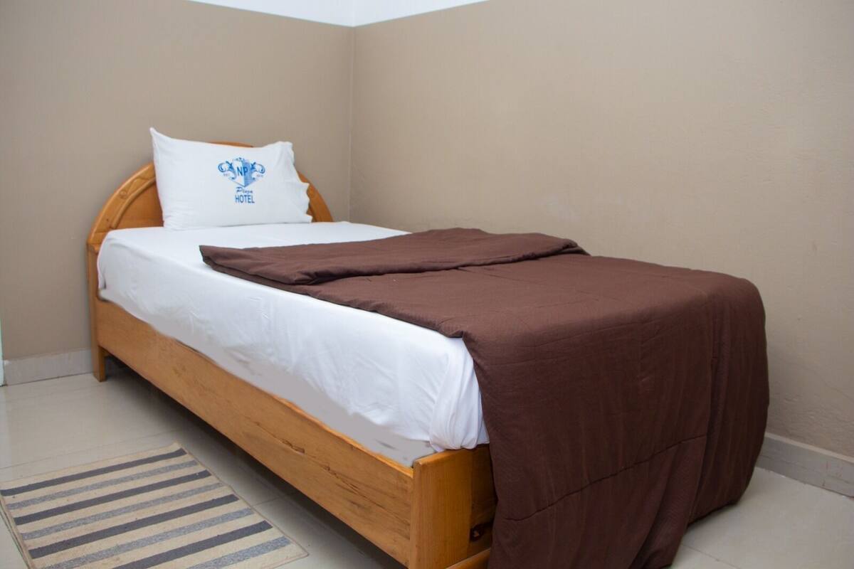 A single bed with a wooden frame, white bedding, a brown blanket, and a pillow with a logo.
