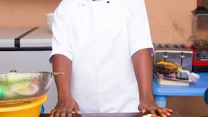 A chef in a white uniform and headscarf standing in a kitchen with two plates of food, one with a fried chicken and rice dish and the other with a salad and rice dish. - NP Plaza Hotel (Akosombo)