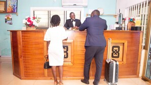 A reception desk with a wooden finish, a glass partition, and a clock on the wall. - NP Plaza Hotel (Akosombo)