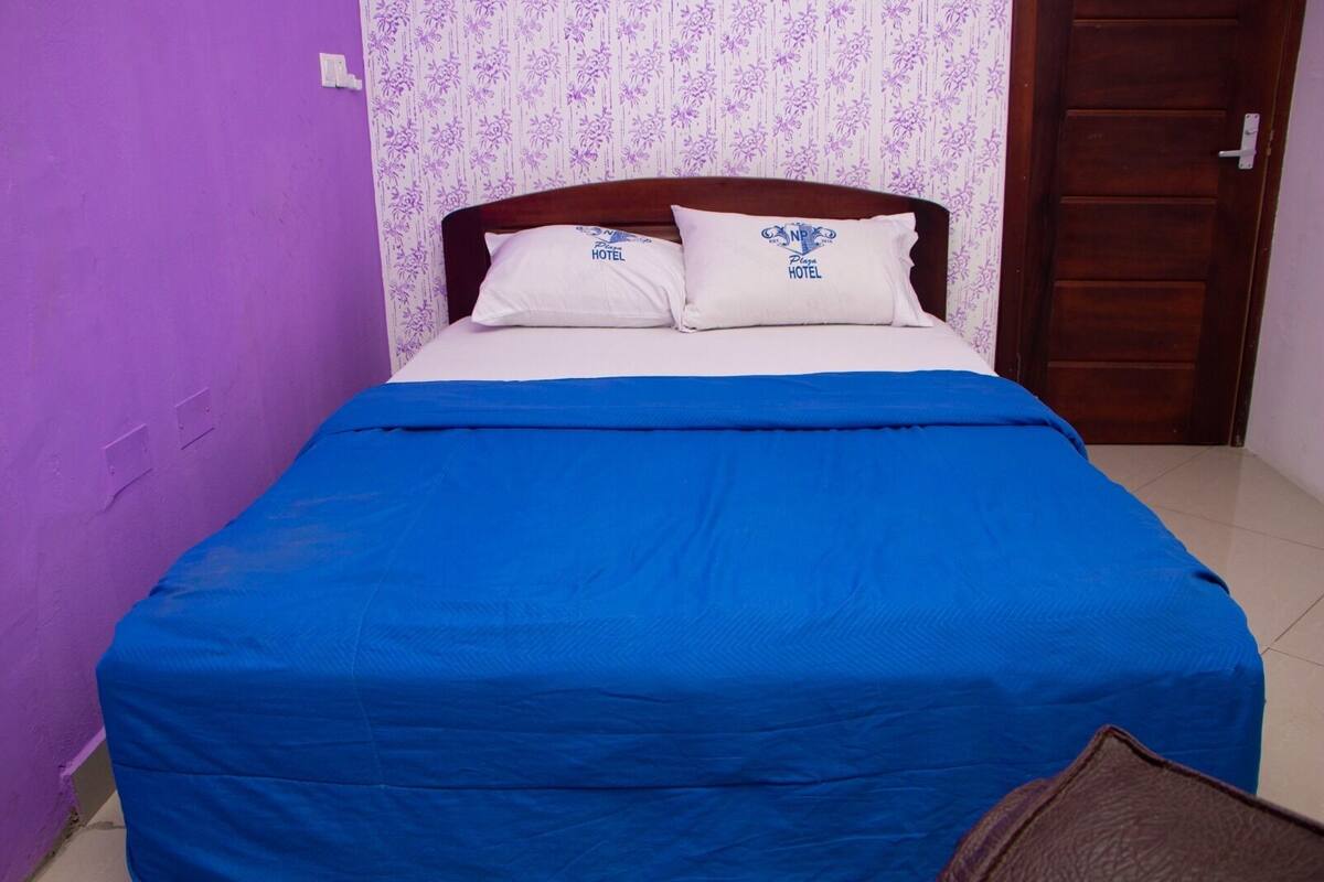 A bed with a blue bedspread and two pillows, a wooden headboard, and a door to the right.