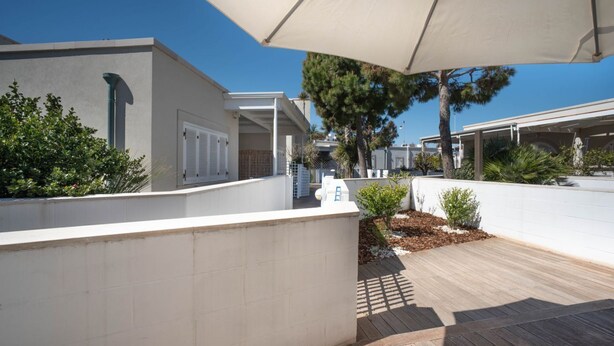 A covered outdoor area with a wooden deck, white walls, and a large umbrella providing shade. - WePuglia - Residence Cala Paura - Apt. Vecchio Frack (Polignano A Mare)