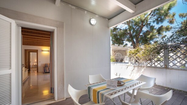 En overdækket udendørs terrasse med et bord og stole, et skilt med teksten 'WePuglia', og udsigt til indretningen gennem en åben dør.