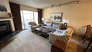 A living room with a fireplace, a sofa, two armchairs, a coffee table, and a large window with curtains.