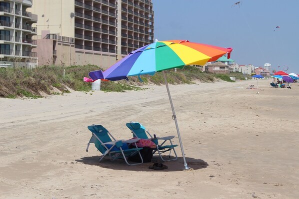 Una spiaggia con un ombrellone colorato, due sedie da spiaggia blu e un'area delimitata con sacchi di sabbia.