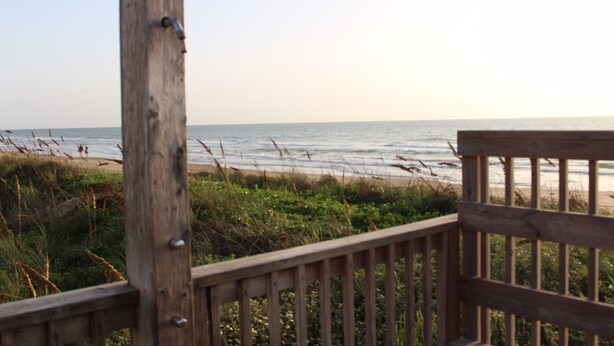 Une terrasse en bois donnant sur une plage, avec vue sur l’océan et des personnes aperçues au loin.