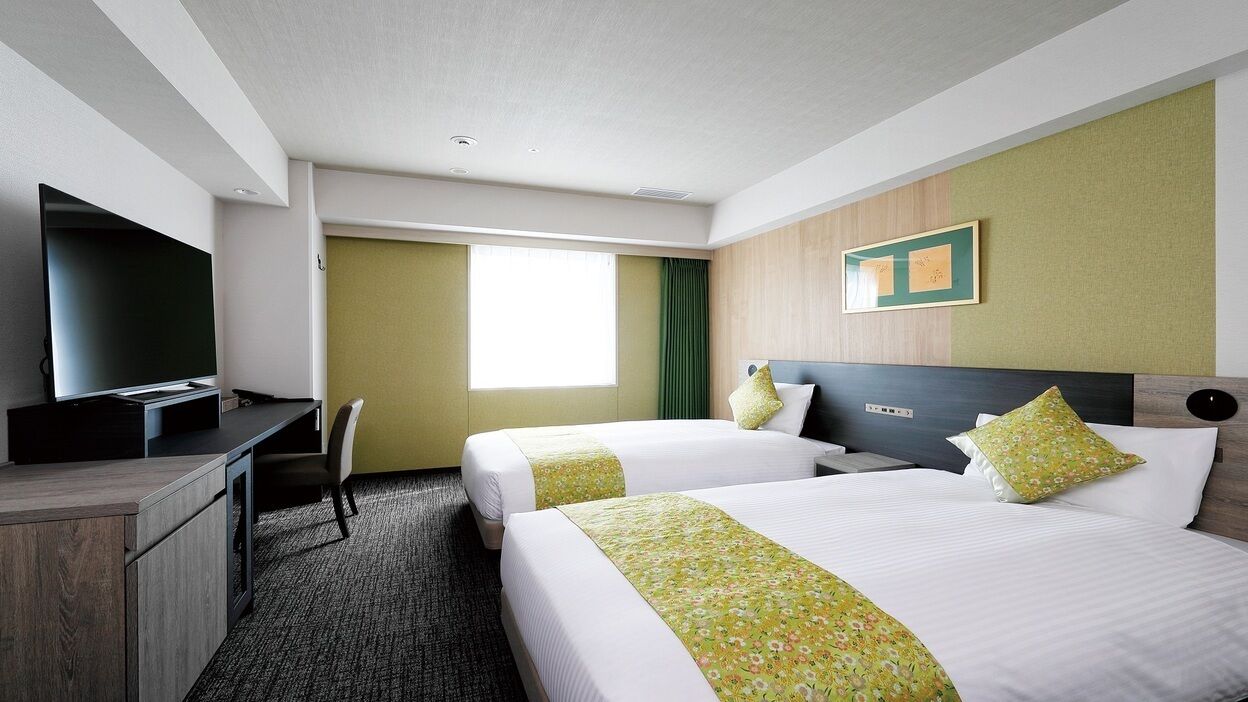 A hotel room with two beds, a desk, a TV, and a window.