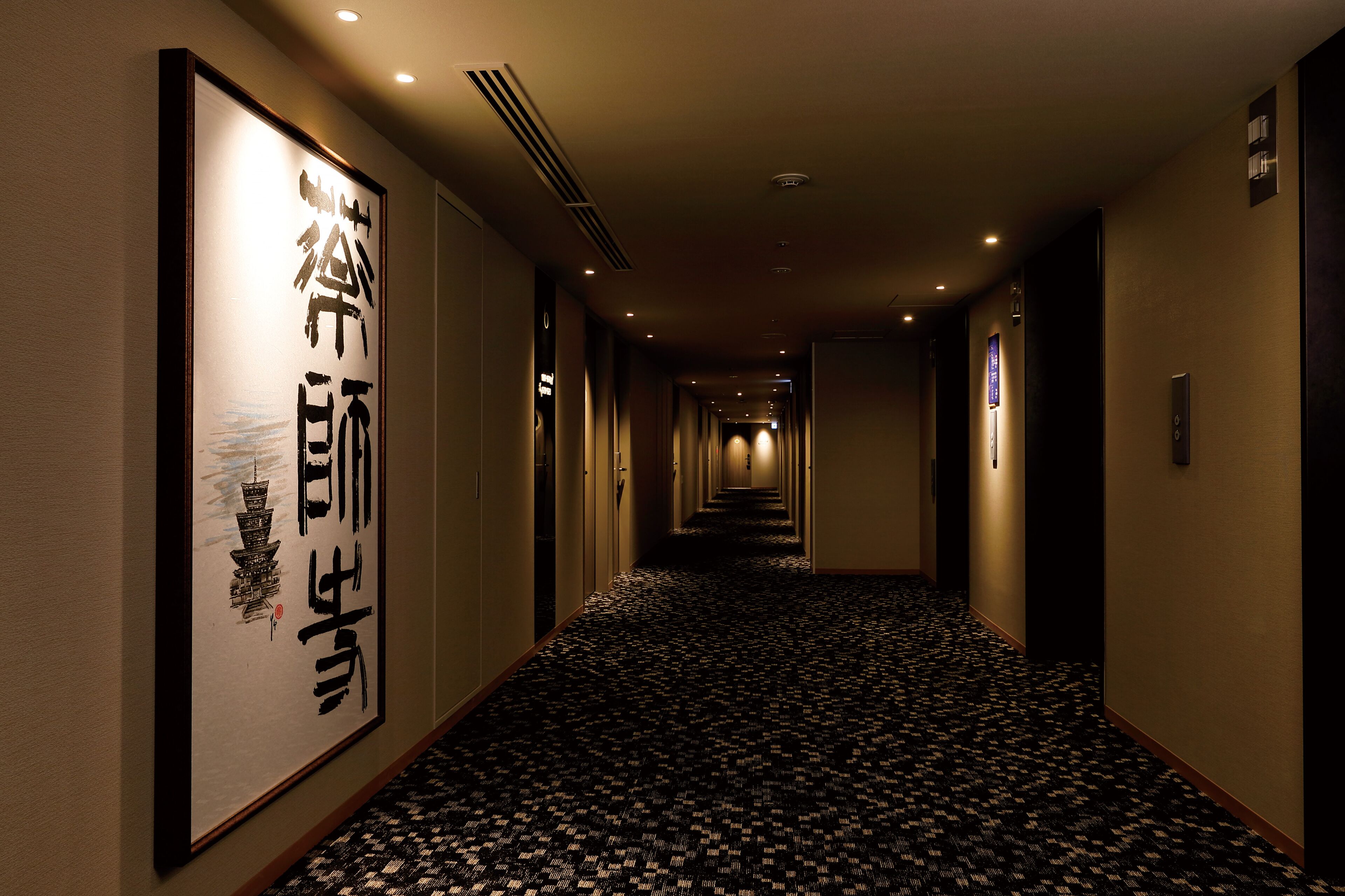 A hotel hallway with a patterned carpet, framed artwork on the walls, and a series of doors leading to rooms.