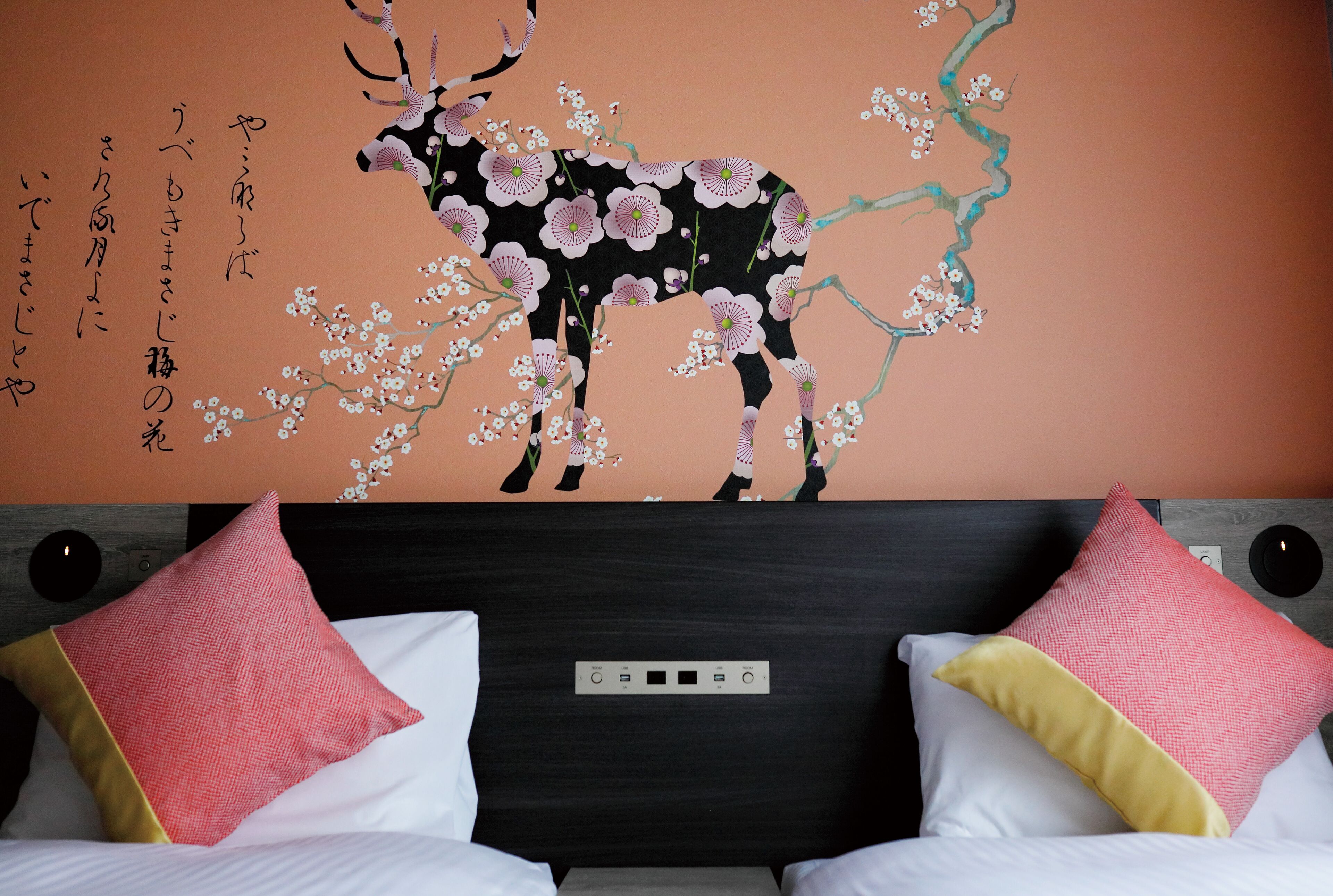 Ein Hotelzimmer mit einem Bett, zwei Kissen, einem Kopfteil mit Bedienfeld und einer Wand mit einer Kirschblüten- und Hirsch-Mural.