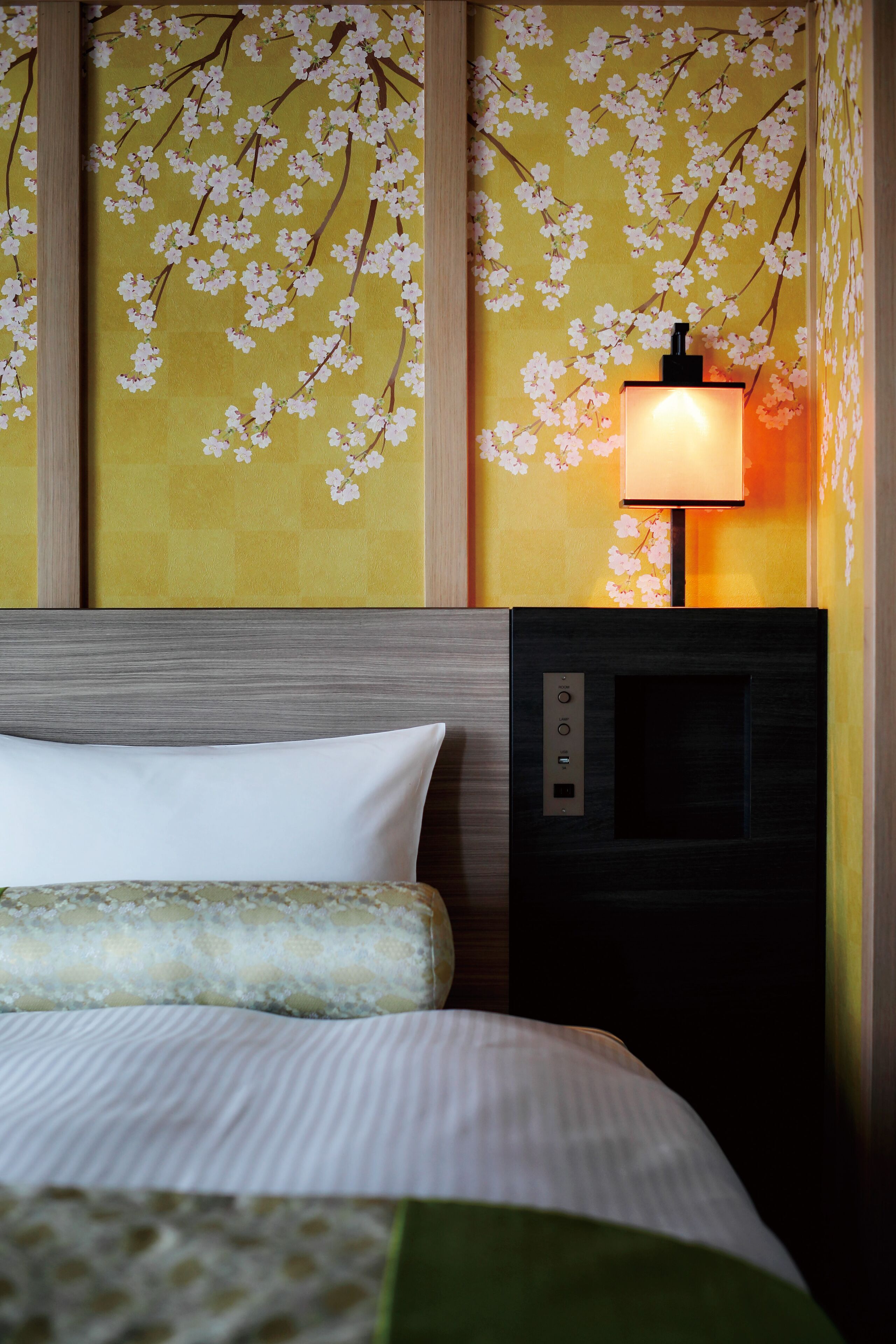 A hotel room with a bed, a nightstand with a lamp, and a wall with cherry blossom wallpaper.