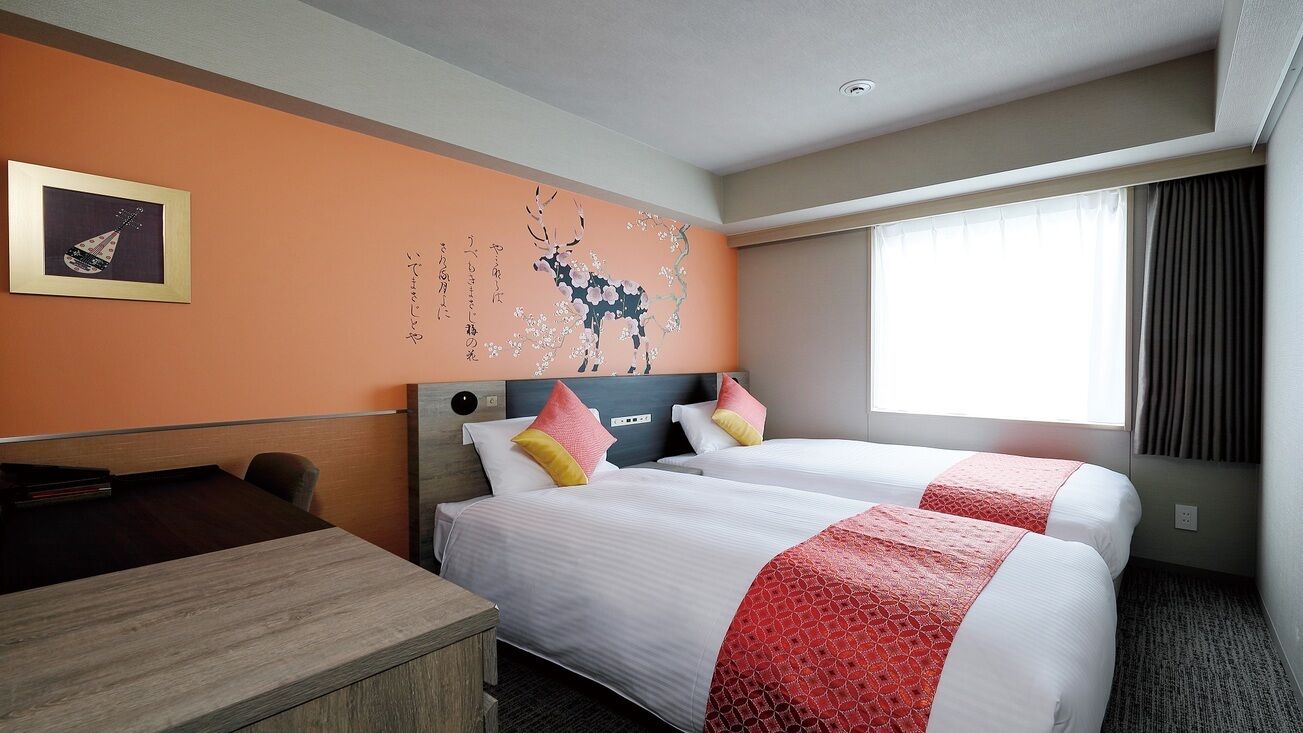 A hotel room with two beds, a desk, a wall with a floral mural, and a large window with curtains.