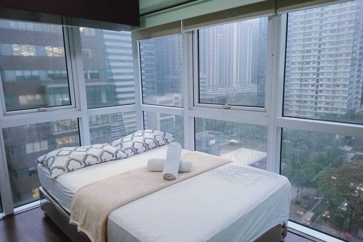 Ein Schlafzimmer mit Bett, Kissen und Blick auf eine Stadtlandschaft durch große Fenster.