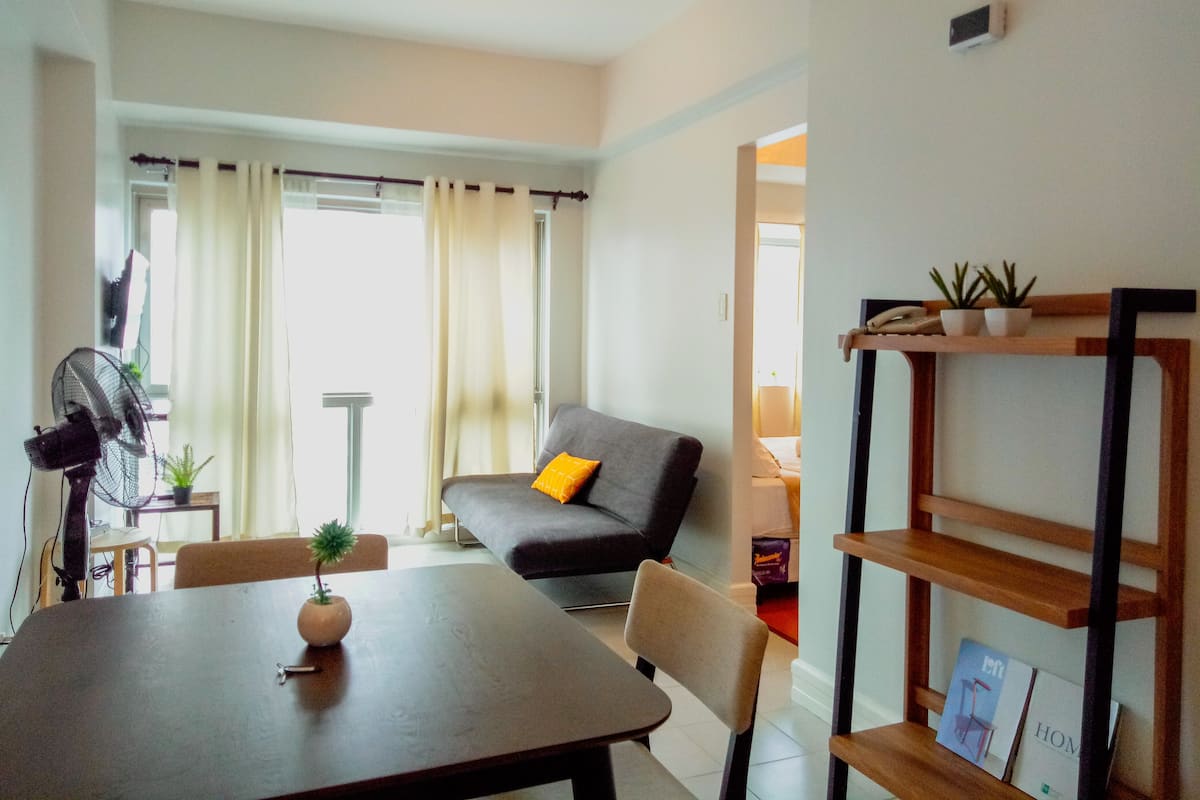 Ein modernes Wohnzimmer mit Esstisch, einer Couch und einem Fernseher. Ücht Blumentöpfe und ein Ventilator sind ebenfalls vorhanden.