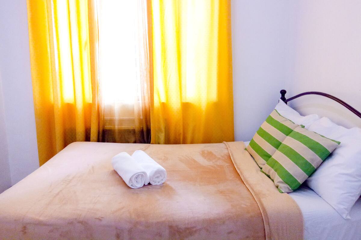 Ein Bett mit gerolltem Handtuch, einem grün-weiß gestreiften Kopfkissen und gelben Vorhängen.