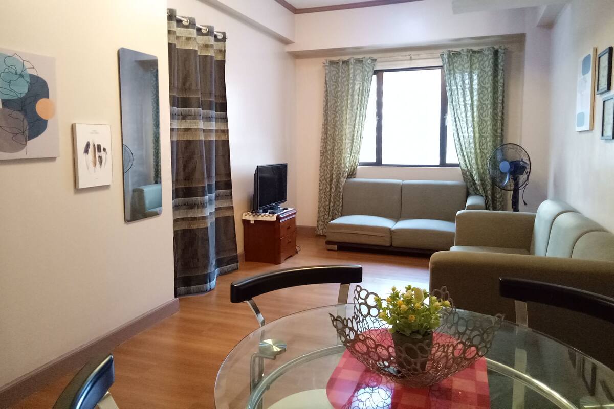 Ein Wohnzimmer mit einer Couch, einem Fernseher, einem Glastisch mit einer Vase und einem Ventilator.