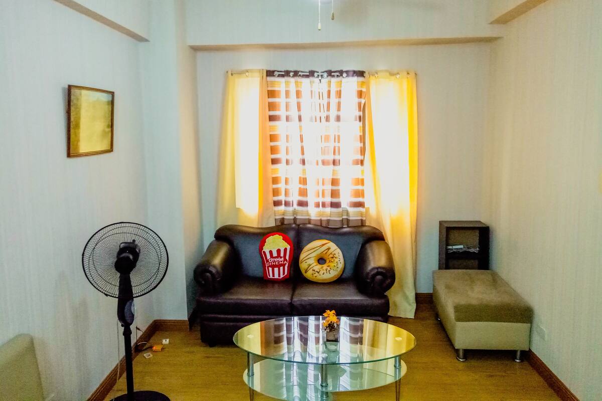 Ein Wohnzimmer mit einer Couch, einem Glastisch, einem Ventilator, einem Fernseher und einem Fenster mit Vorhängen.