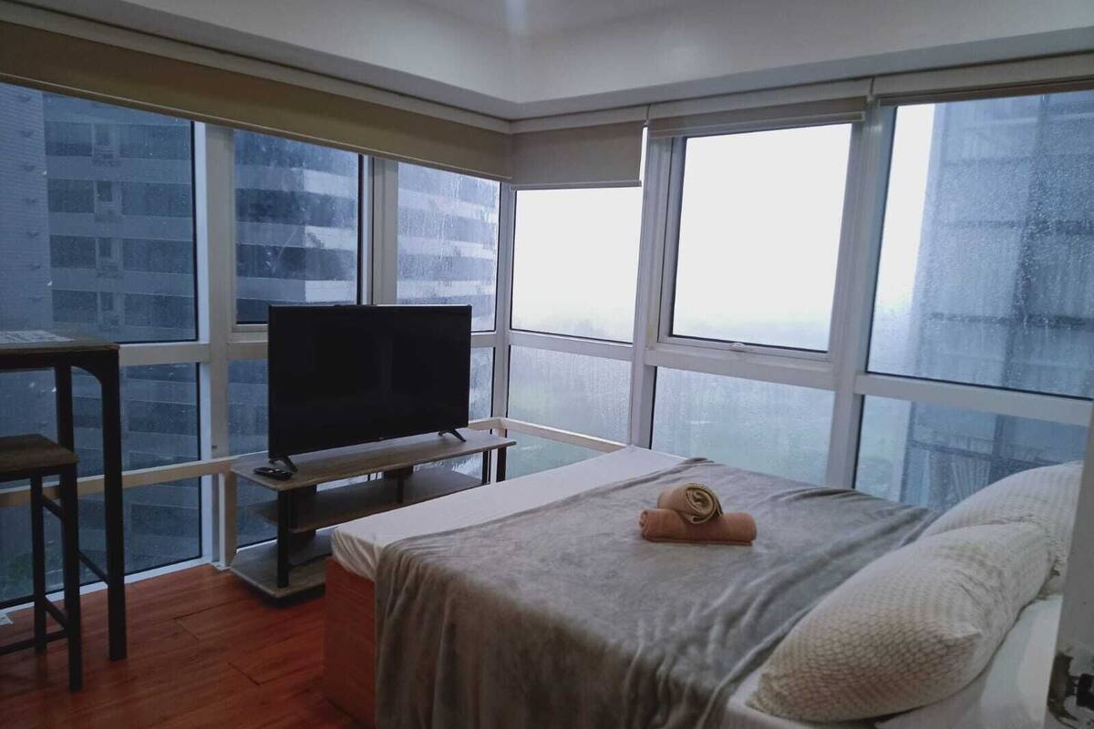 Ein Schlafzimmer mit einem Bett, einem Fernseher auf einem Schrank, einem Schreibtisch und großen Fenstern.