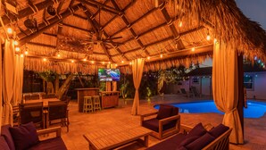 An outdoor lounge area with a thatched roof, wooden furniture, a pool, and a television screen.