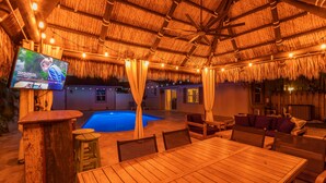 A thatched-roof outdoor area with a pool, wooden furniture, and a television screen.