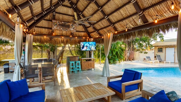 An outdoor lounge area with a thatched roof, wooden furniture, a pool, and a TV screen.