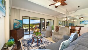 A spacious living room with a large gray sofa, a glass coffee table, and a view of a garden through the sliding glass doors. - SUMMER SPECIAL! Spacious Luxury 2 Bedroom Condo, Maui Sunsets! No Resort Fees! (Lahaina)
