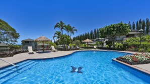 Une piscine à l’eau d’un bleu cristallin, entourée de chaises longues, de palmiers et d’une structure à toit de chaume.