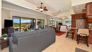 A modern living room with a grey sofa, a flat-screen TV, and a dining area with wooden chairs and a red rug. - SUMMER SPECIAL! Spacious Luxury 2 Bedroom Condo, Maui Sunsets! No Resort Fees! (Lahaina)