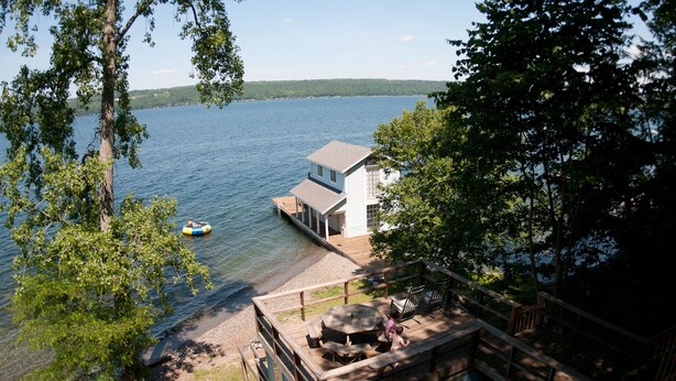 Una cabaña junto al lago con muelle, un bote pequeño y una terraza con mobiliario exterior.