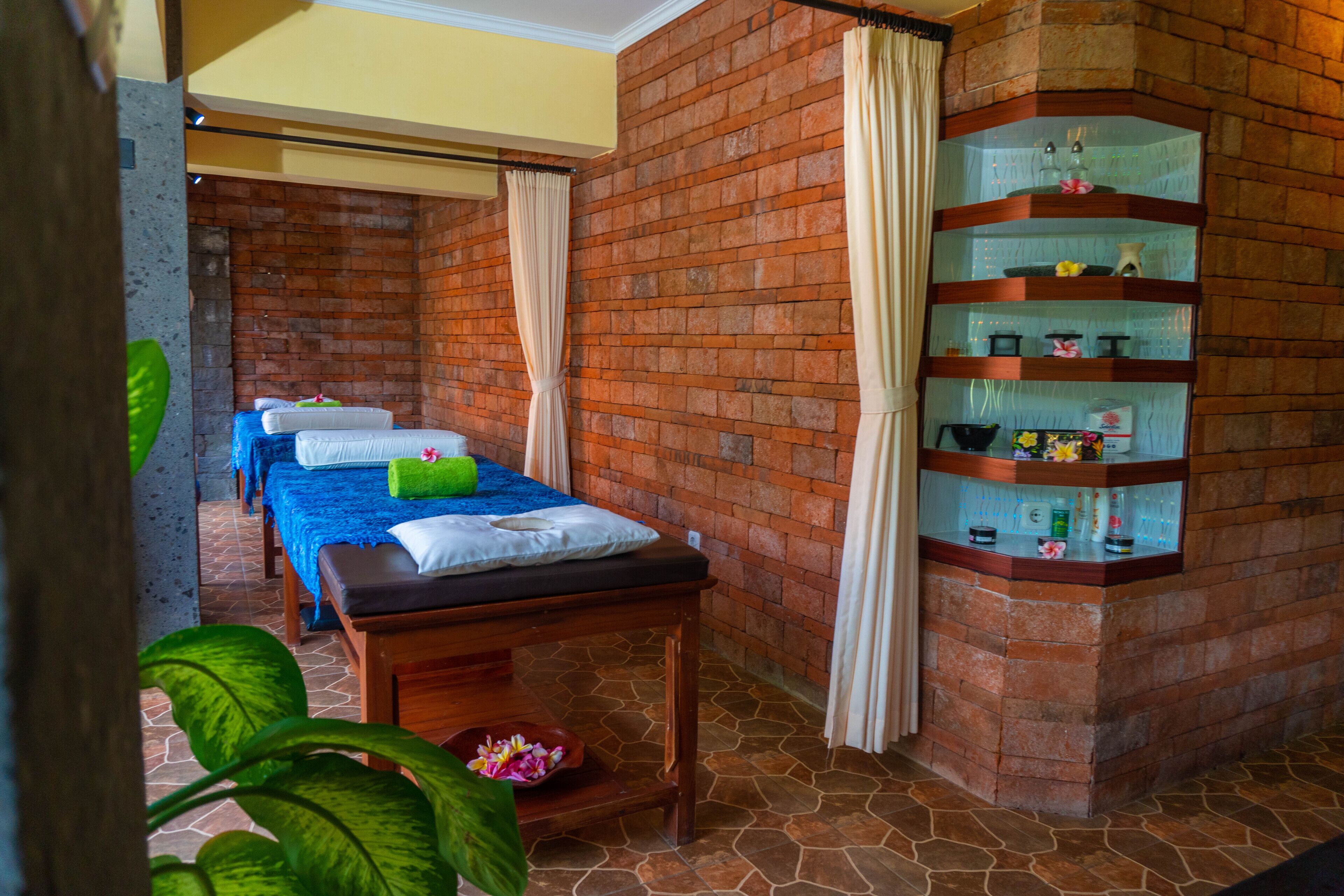 A spa room with a massage table, two massage beds, a shelf with toiletries, and a brick wall.