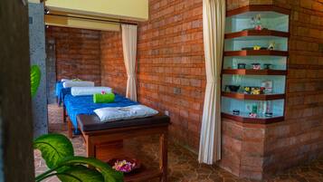 A spa room with a massage table, two massage beds, a shelf with toiletries, and a brick wall.