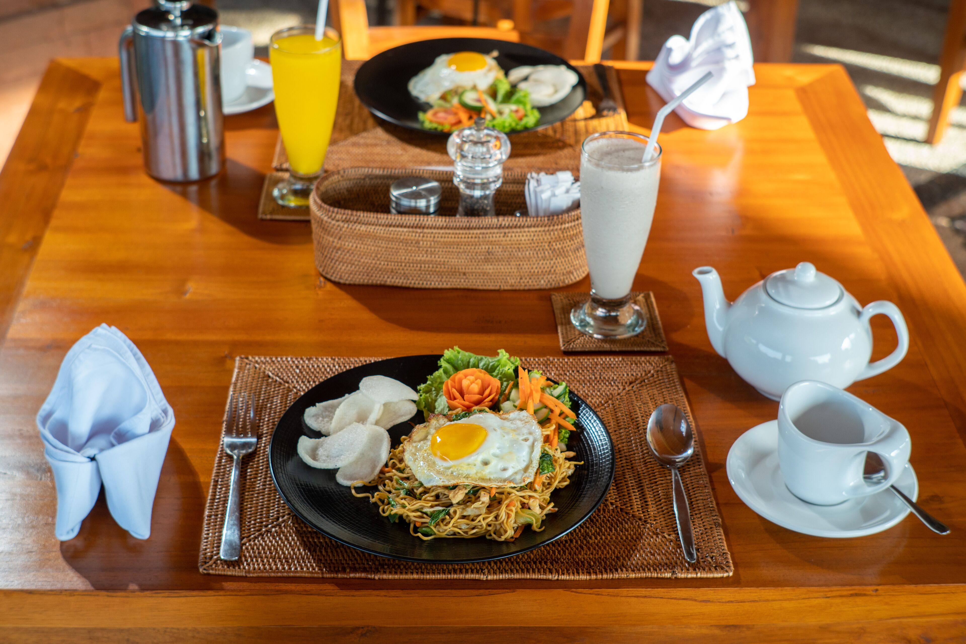 Una mesa de madera con un plato negro de fideos coronado con un huevo frito, una tetera blanca, una taza blanca y un vaso de leche.