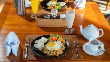 Una mesa de madera con un plato negro de fideos coronado con un huevo frito, una tetera blanca, una taza blanca y un vaso de leche.
