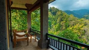 A balcony with a wooden bench, a stone floor, and a view of a lush forest and mountains.