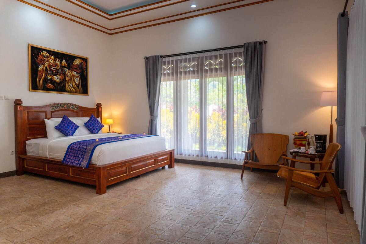 A bedroom with a large bed, a wooden headboard, a nightstand, a chair, a table, and a window with curtains.