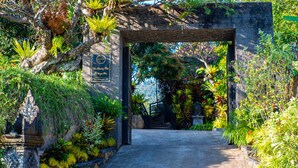 A stone archway leading into a lush garden with various plants and a small Buddha statue.